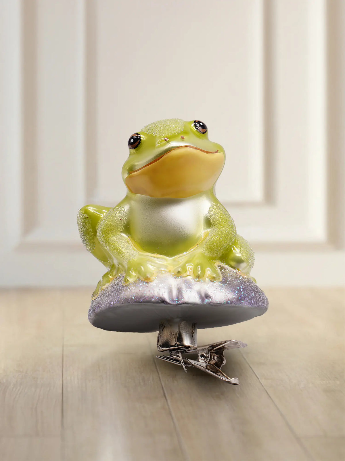 Glasornament Frosch: hellgrüner sitzender Frosch mit gelber Kehle und Glitzer auf fliederfarbenem Sockel, mit silbernem Clip zum Anklemmen