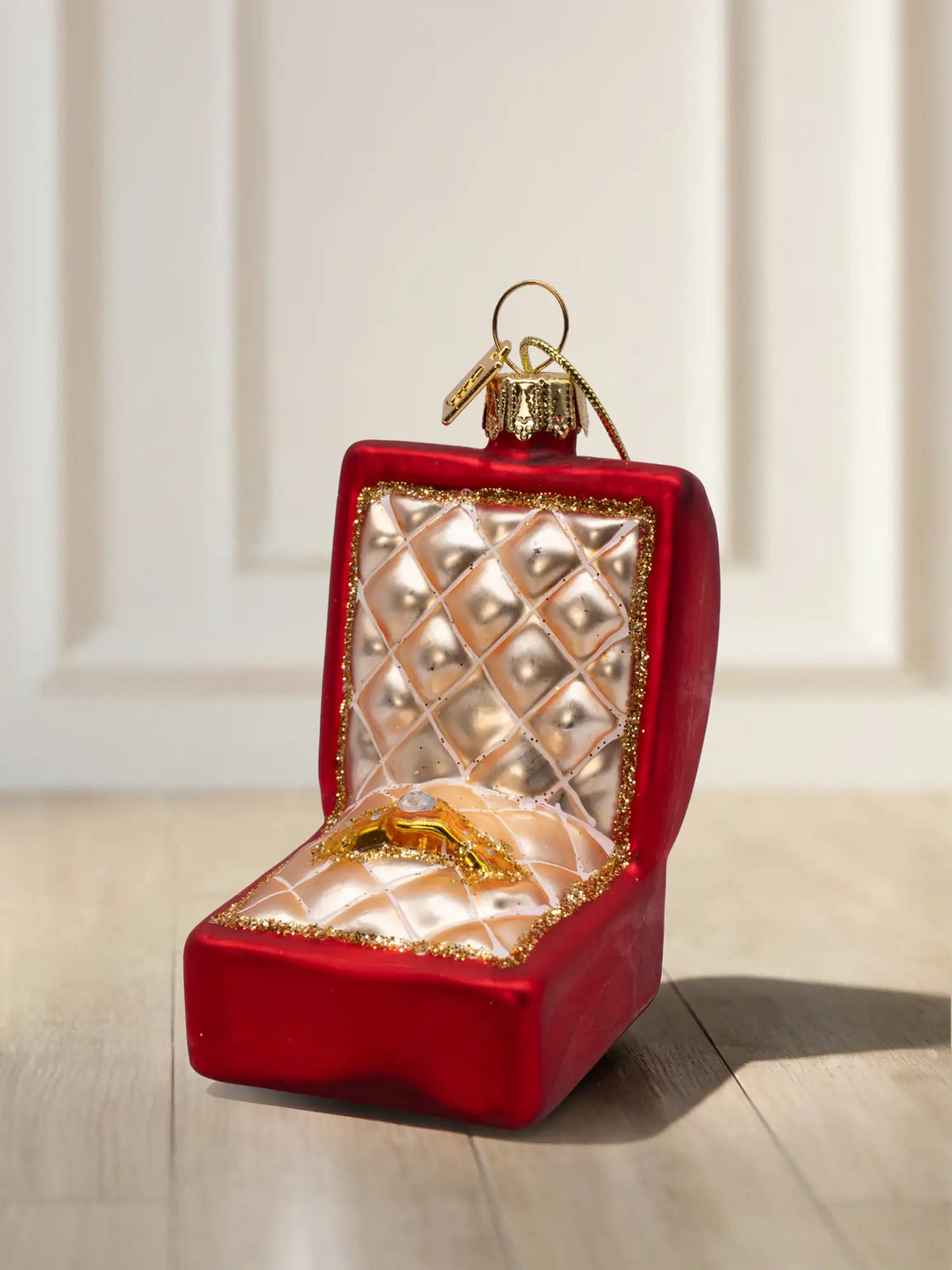 Rotes, mattes Glasornament in Form einer geöffneten Ringschatulle mit goldgesteppter Polsterung, goldenem Ring mit Stein und Goldglitzerrand.