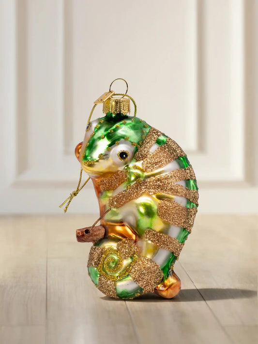 Glasornament in Form eines Chamäleons mit grün-weißem Körper, goldenen Glitzerstreifen, eingerolltem Schwanz und einem Ast, auf Holzboden.