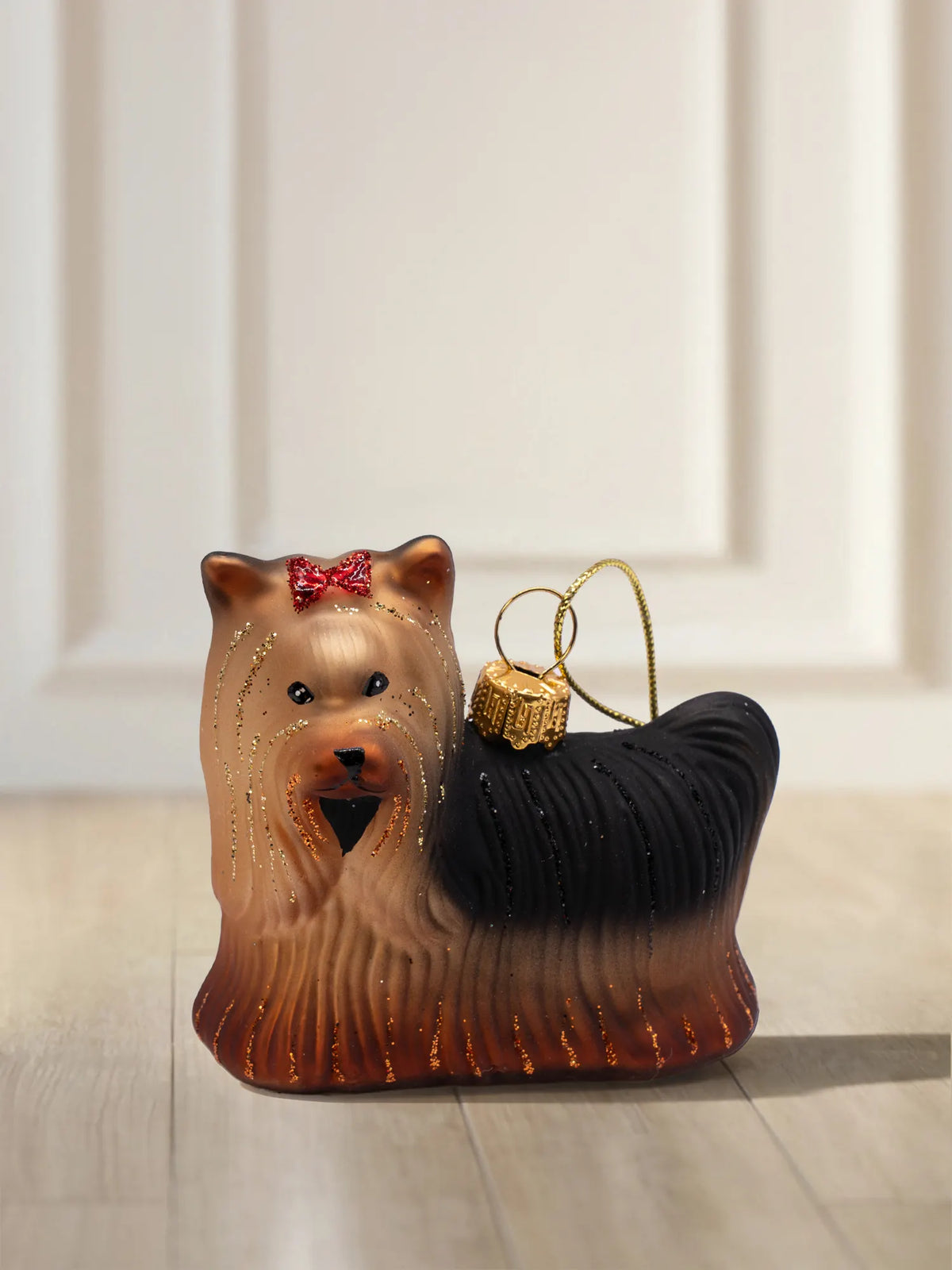 Glasornament in Form eines Yorkshire Terriers mit rotem Glitzerschleifchen im Haar, schwarzem Rücken, goldbraunem Fell und feinen Glitzerlinien.