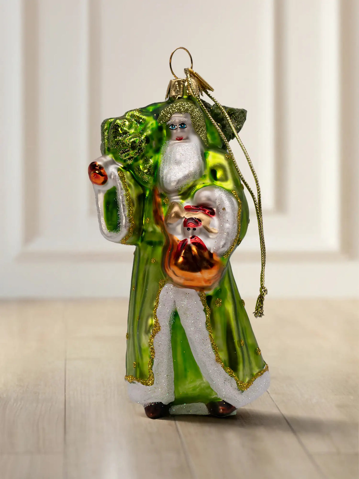 Glasornament Waldsanta: Santa in langem grünem Mantel mit weißem Fellbesatz und goldenen Glitzerdetails, hält einen kleinen Weihnachtsbaum, goldene Aufhängekappe
