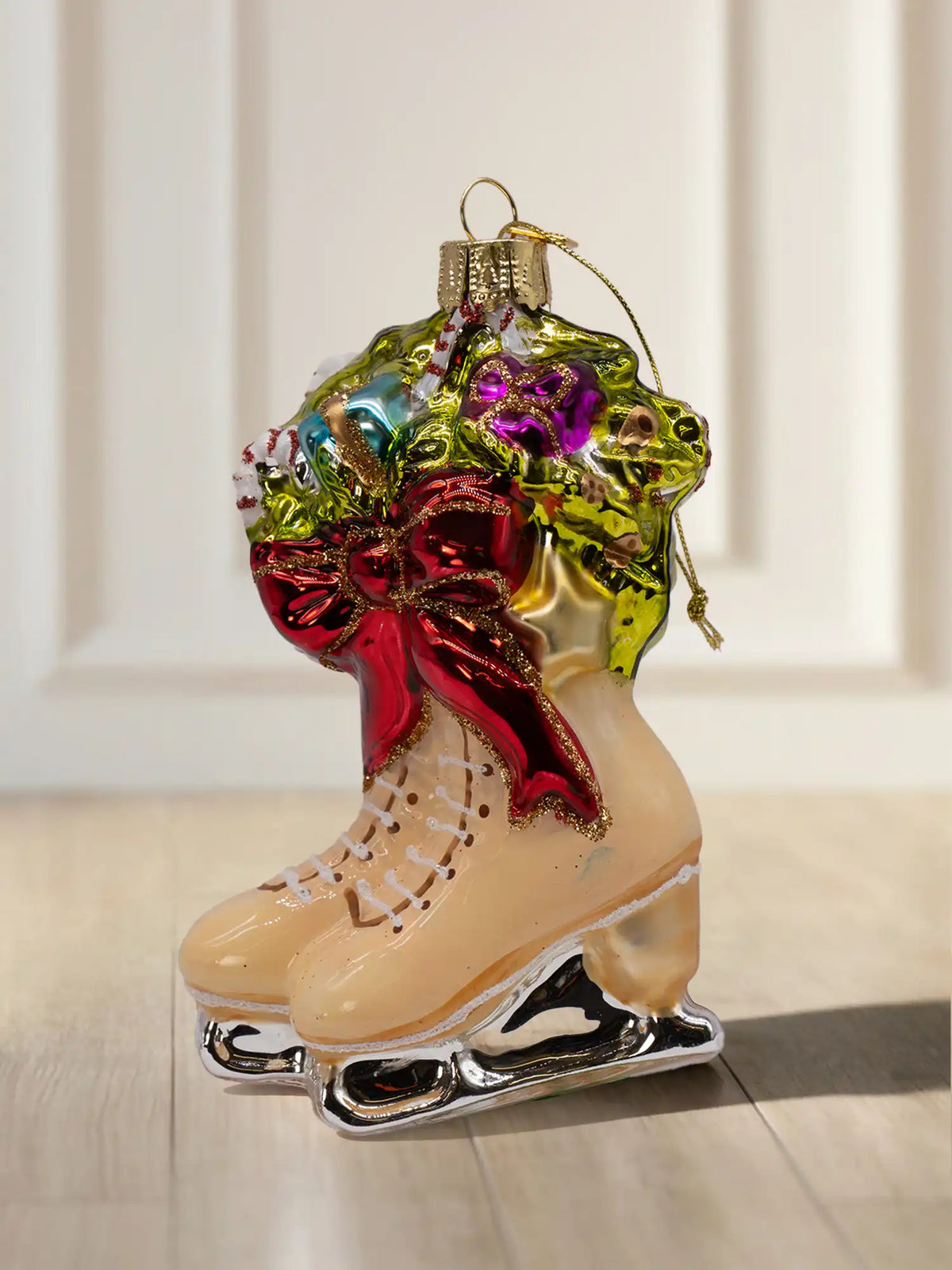 Glasornament in Form eines Paars Schlittschuhe in Creme mit silbernen Kufen, roter Schleife und einem Gesteck aus Tannengrün, Zuckerstangen und Geschenken.