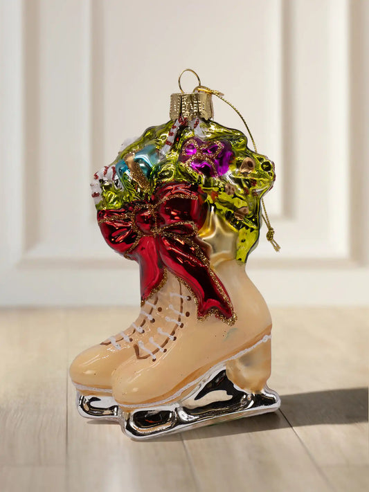 Glasornament in Form eines Paars Schlittschuhe in Creme mit silbernen Kufen, roter Schleife und einem Gesteck aus Tannengrün, Zuckerstangen und Geschenken.