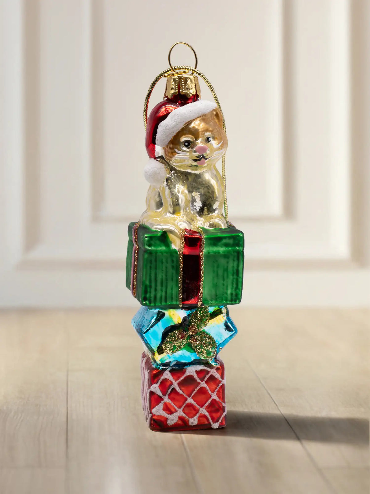 Glasornament Katze auf Geschenk: Goldbraune Katze mit roter Weihnachtsmütze sitzt auf einem Stapel aus drei Geschenkboxen in Grün, Blau und Rot mit Glitzerband, goldene Aufhängekappe