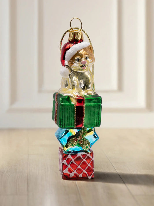 Glasornament Katze auf Geschenk: Goldbraune Katze mit roter Weihnachtsmütze sitzt auf einem Stapel aus drei Geschenkboxen in Grün, Blau und Rot mit Glitzerband, goldene Aufhängekappe
