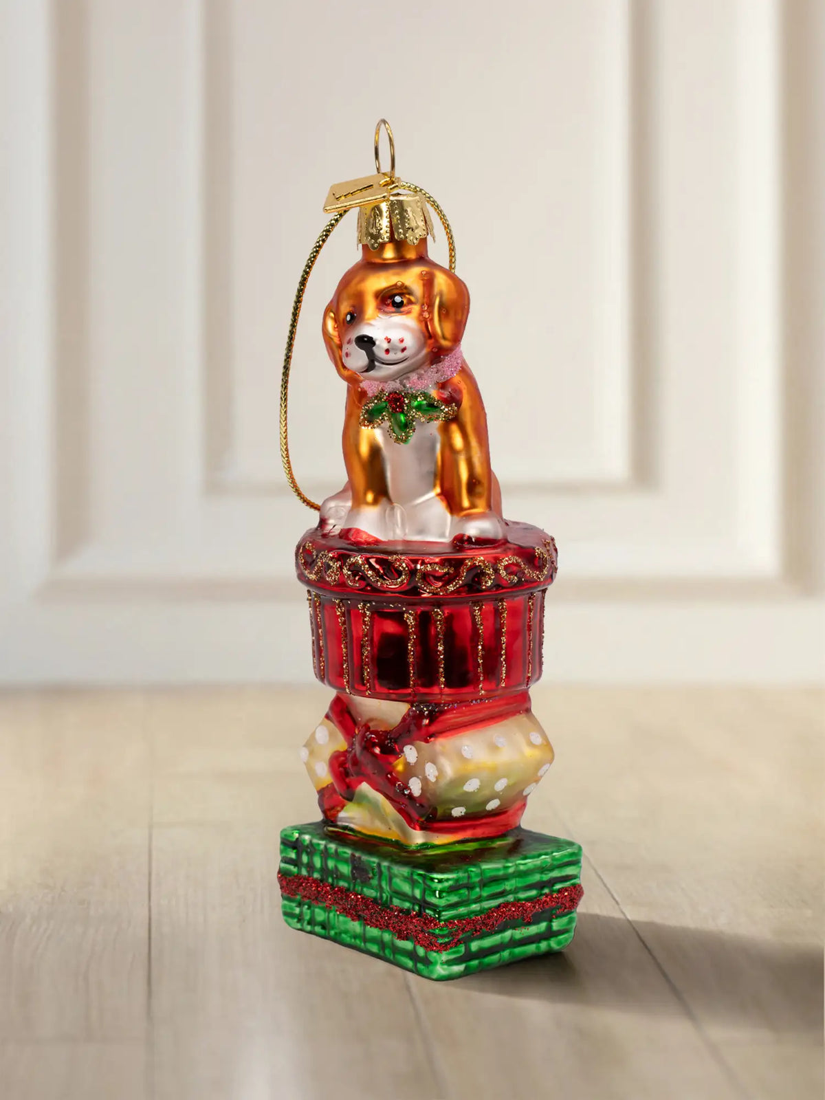 Glasornament Hund auf Geschenk: brauner Welpe sitzend auf gestapelten Weihnachtspäckchen (roter Korb, gepunktetes Päckchen, grüne Box), Stechpalme am Halsband, goldenes Hängeelement