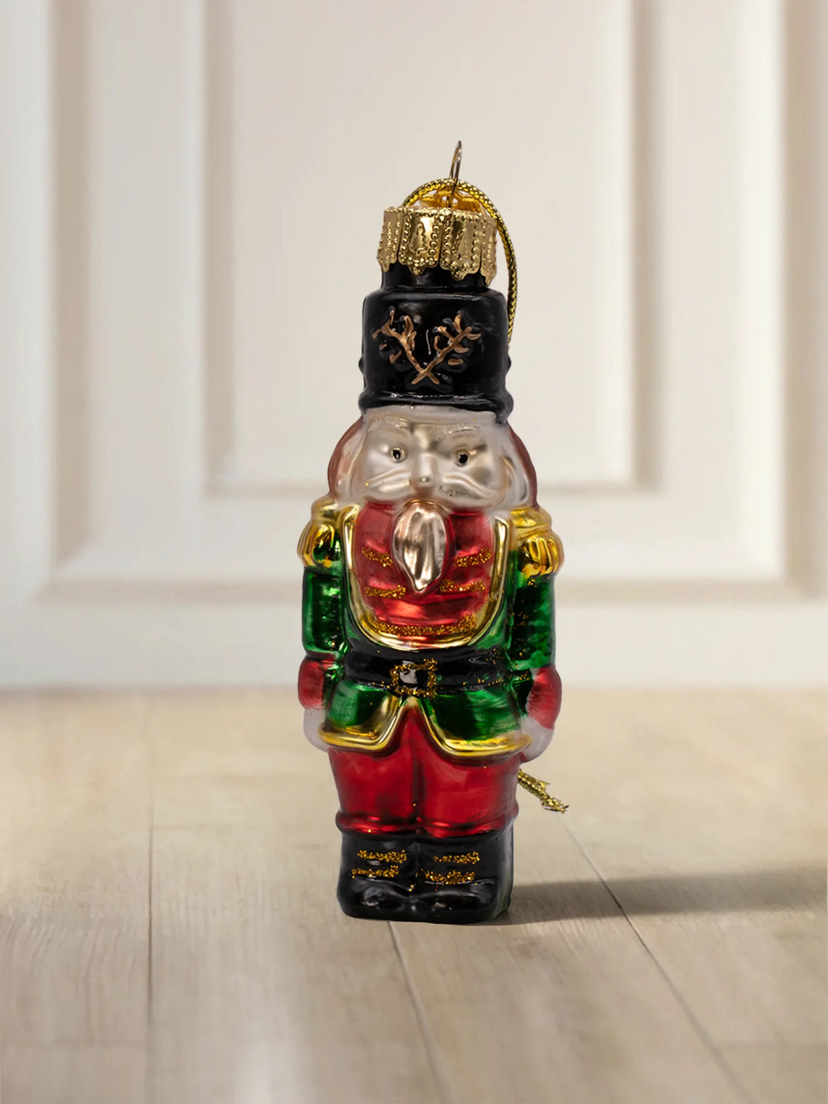 Glasornament Nussknacker: Roter Uniformrock mit grünen Ärmeln und goldenen Schulterdetails, schwarze Mütze mit goldenem Lorbeermotiv, weißer Bart, goldene Aufhängekappe