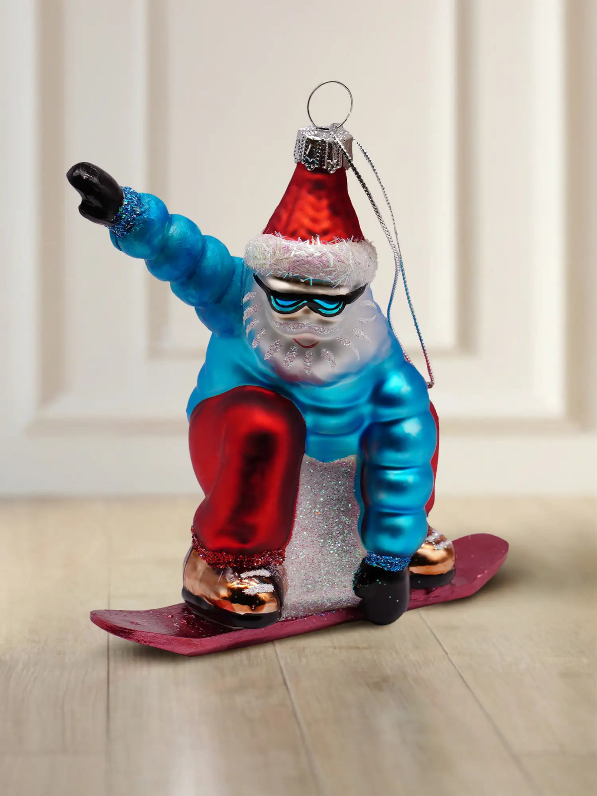Glasornament Santa auf Snowboard: Weihnachtsmann in türkisblauem Schneeanzug mit roter Mütze und weißem Fellrand, türkisfarbene Skibrille, rotes Snowboard mit goldenen Bindungen, silberne Aufhängekappe