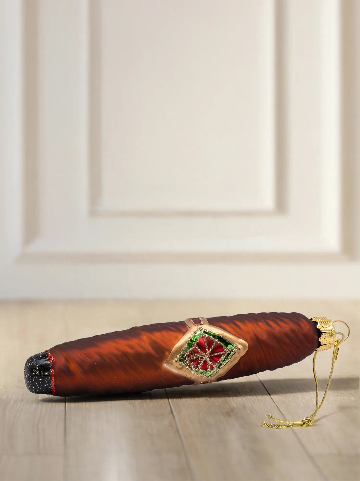 Braunes Glasornament in Form einer Zigarre mit goldener Bauchbinde in Rot und Grün, schwarz glitzerndem Ende und goldener Aufhängung, auf Holzboden liegend.
