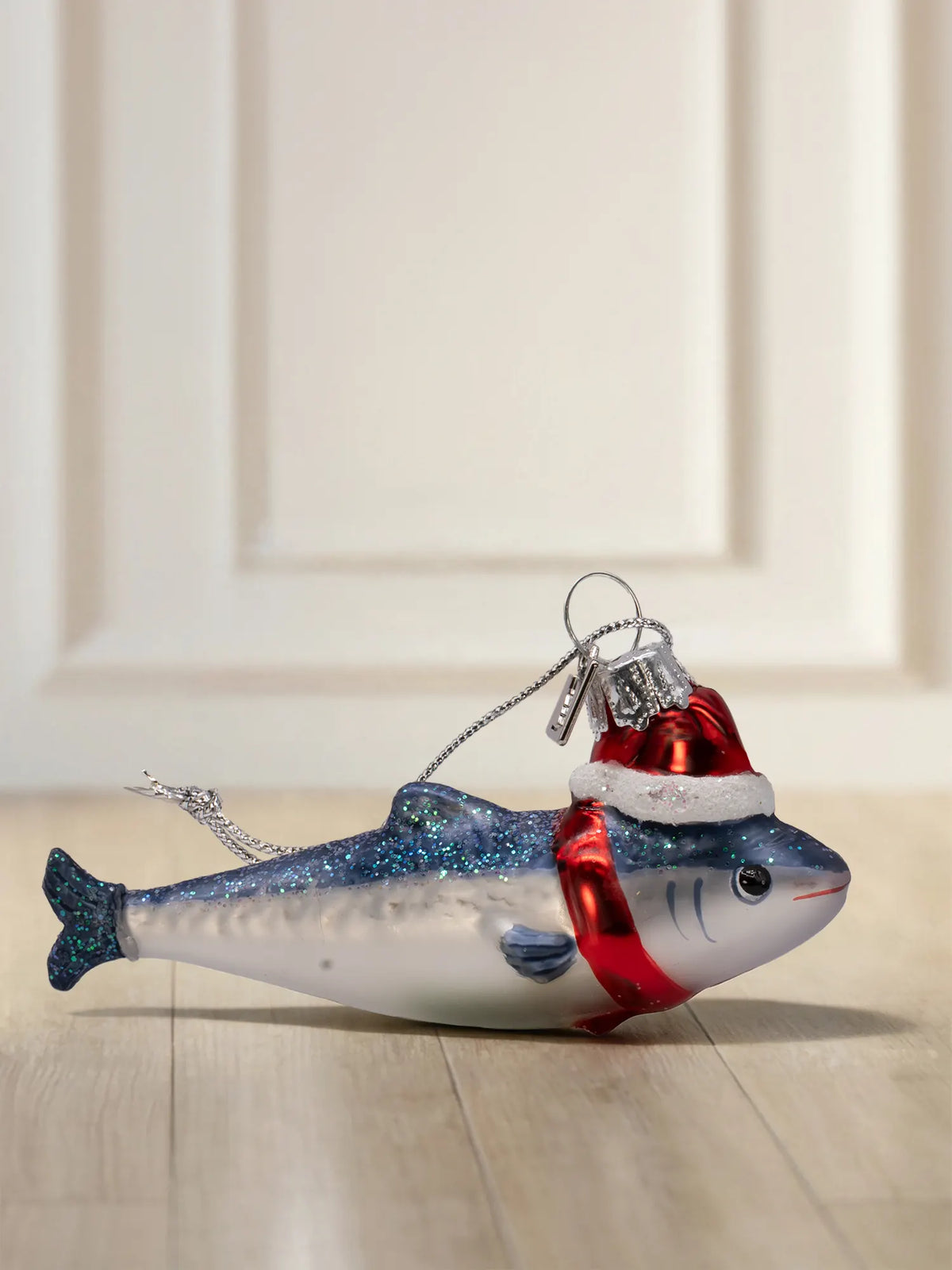 Glasornament in Form eines blauen Fisches mit Glitzerrücken, roter Weihnachtsmütze und rotem Schal, mit silberner Aufhängung.