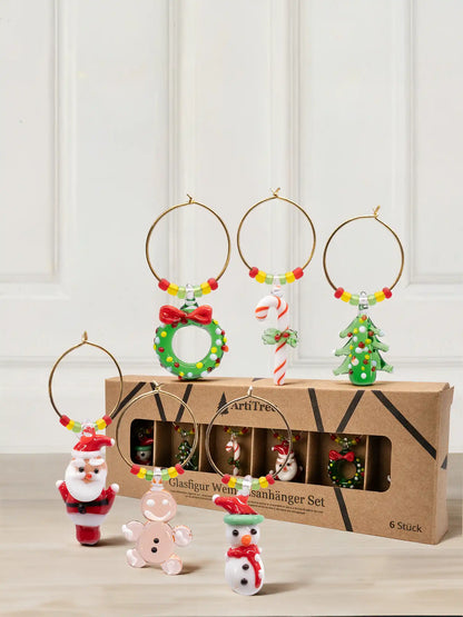 Set aus sechs Weinglasanhängern mit goldfarbenen Ringen und bunten Perlen: Weihnachtsmann, Lebkuchenmännchen, Schneemann, Kranz, Zuckerstange und Tannenbaum aus Glas vor der Kartonverpackung mit Sichtfenster.