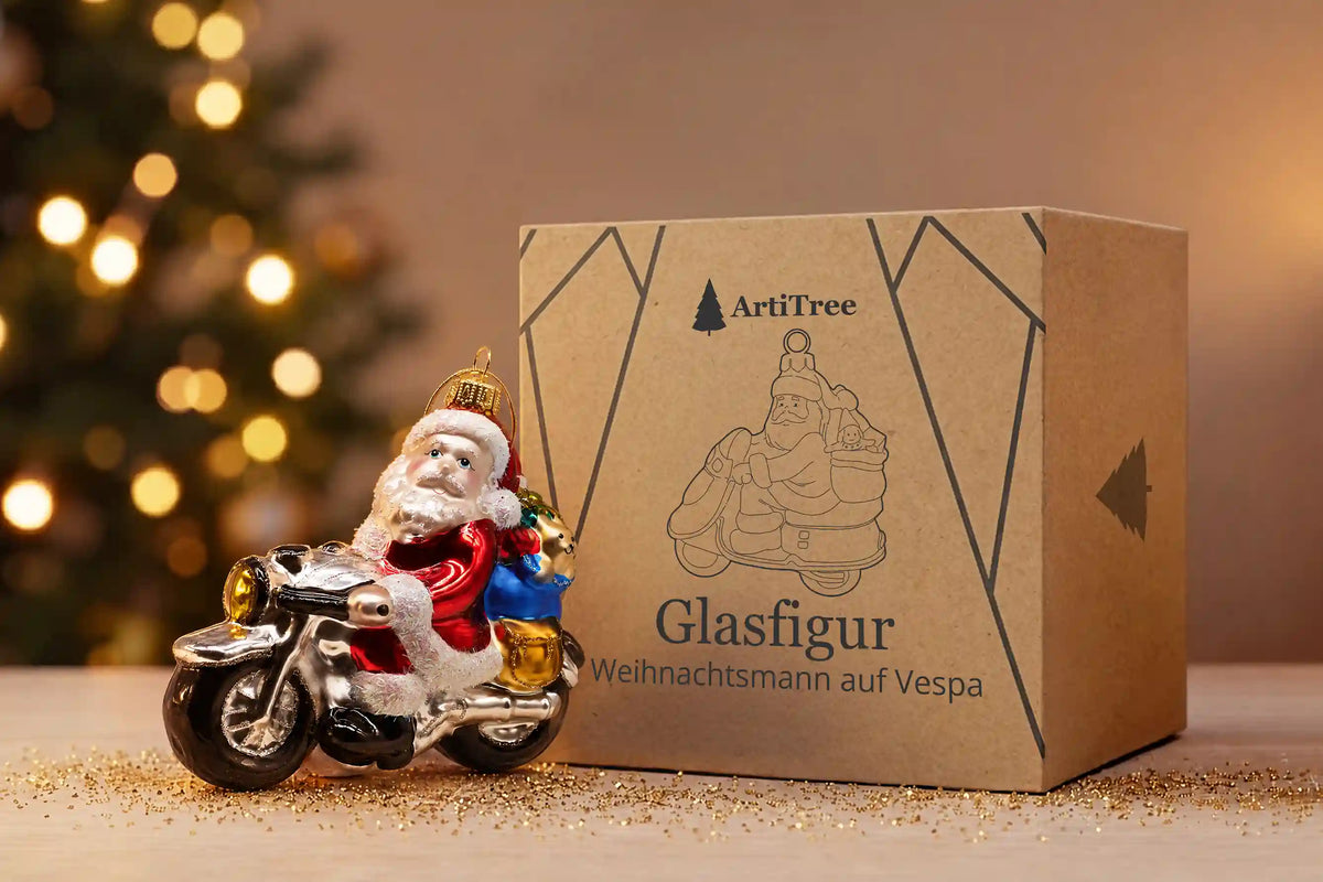 Glasornament Weihnachtsmann auf Zweirad mit Geschenkesack neben der ArtiTree-Verpackung aus Pappe mit Aufschrift „Glasfigur Weihnachtsmann auf Vespa“, im Hintergrund ein beleuchteter Weihnachtsbaum