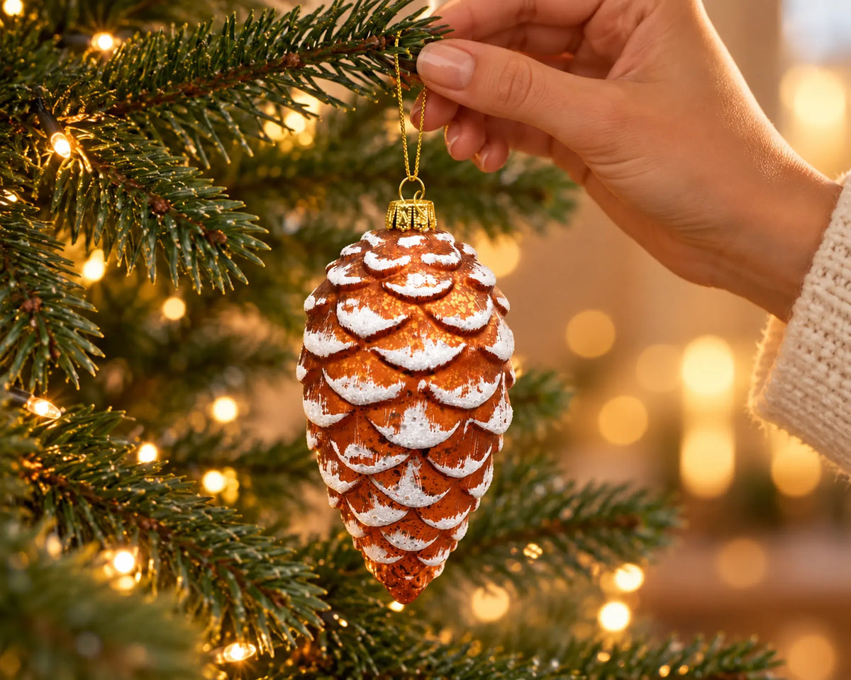 Hand hängt eine Glasfigur in Form eines Tannenzapfens an einen beleuchteten Weihnachtsbaum, kupferbraun mit weiß verschneiten Schuppen und goldener Kordel