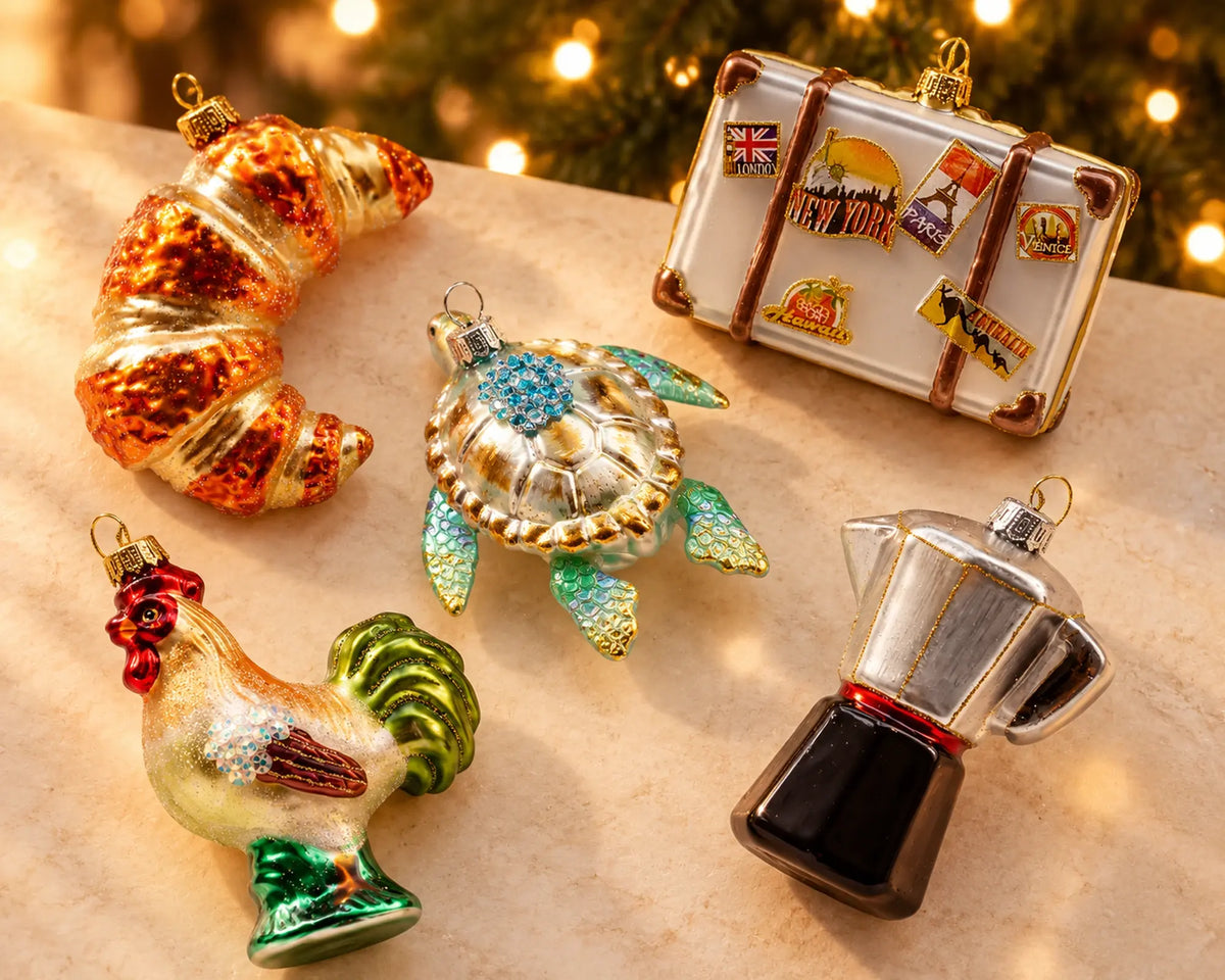 Fünf Glasfiguren auf heller Steinfläche vor Lichterkette: Croissant, Meeresschildkröte, Reisekoffer mit Aufklebern, Hahn und Espressokocher