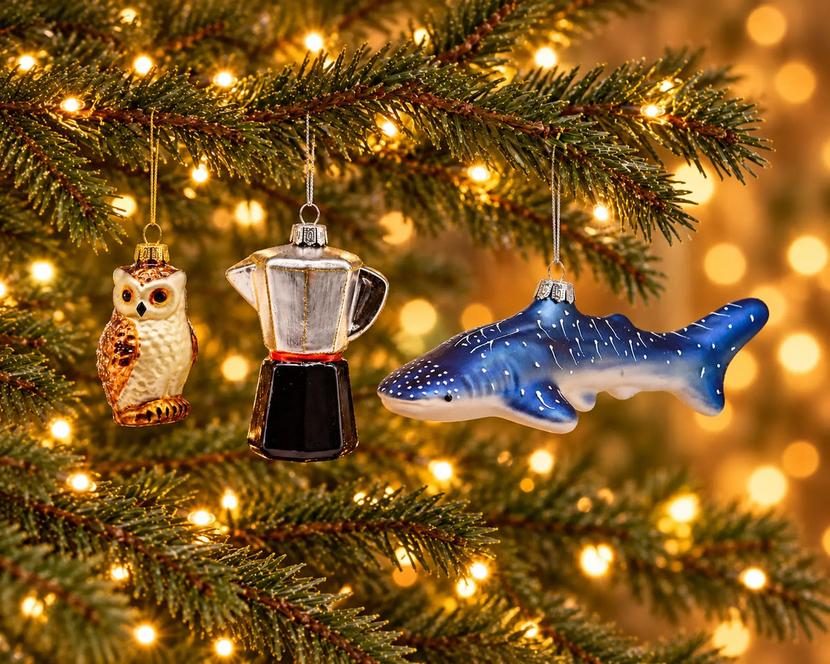 Drei Glasfiguren hängen nebeneinander am beleuchteten Weihnachtsbaum im Größenvergleich: kleine Eule, mittelgroßer Espressokocher und langer Walhai